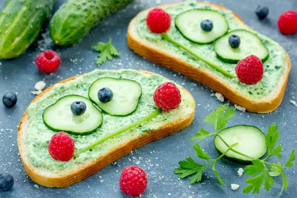 Gurkenscheiben, Petersilienstängel und Himbeeren bilden Gesichter auf belegten Brotscheiben.