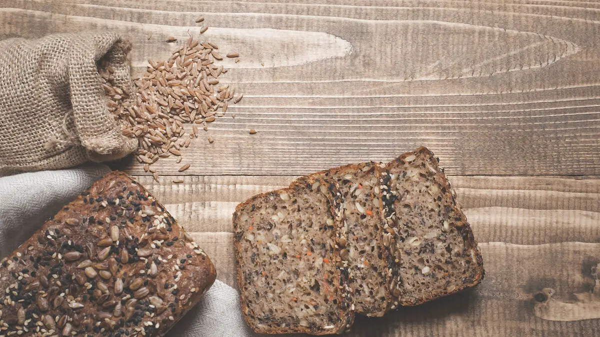 Zwei Scheiben Körnerbrot liegen auf einem Holzbrett, darüber ein Sack mit Körnern.