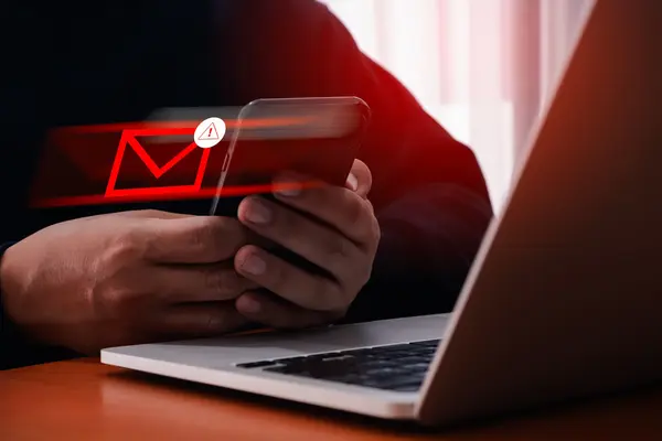 Nahaufnahme Laptop, davor Hände mit Smartphone. Daraus fliegt ein roter Briefumschlag: Phishing Amazon