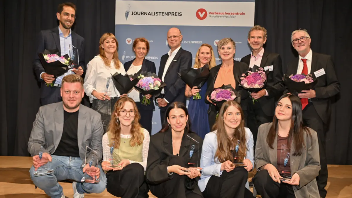 Gruppenbild von 13 Personen, die in die Kamera lächeln, stehend und kniend vor einem Aufsteller mit dem Schriftzug "Journalistenpreis" und dem Logo der Verbraucherzentrale Nordrhein-Westfalen