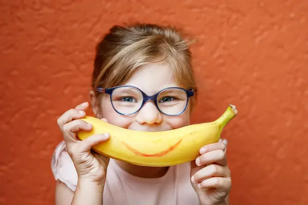 Ein Mädchen mit Brille hält sich einen Banane mit einem aufgemalten Lächeln vor den Mund.