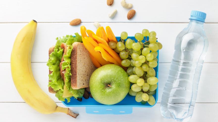 In einer Brotzeitbox liegen ein belegtes Brot, ein grüner Apfel, Paprika-Sticks und grüne Weintrauben. Links daneben liegt eine Banane. Rechts neben der Box liegt eine Flasche mit Wasser.