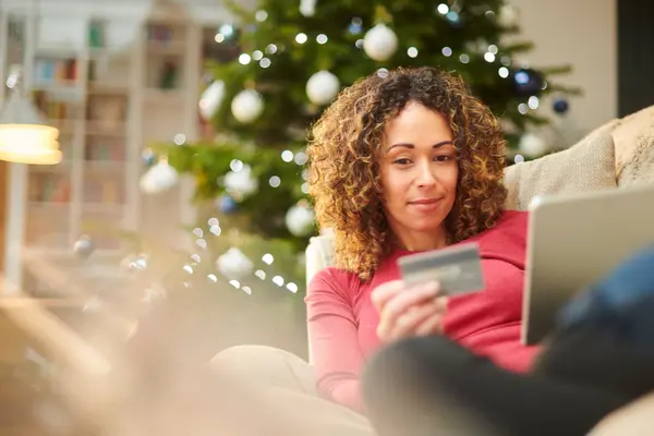 junge Frau auf dem Sofa mit Laptop und Kreditkarte, im Hintergrund ein Weihnachtsbaum