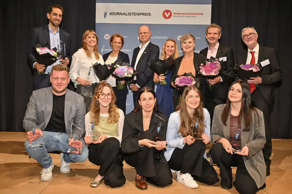 Gruppenbild von 13 Personen, die in die Kamera lächeln, stehend und kniend vor einem Aufsteller mit dem Schriftzug "Journalistenpreis" und dem Logo der Verbraucherzentrale Nordrhein-Westfalen