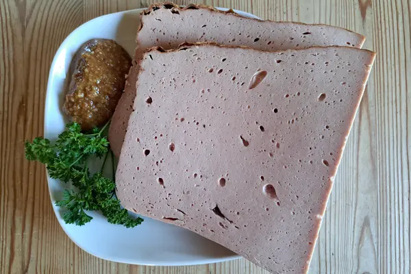 Zwei Scheiben Leberkäse liegen auf einem weißen Teller; daneben süßer Senf un Petersilie