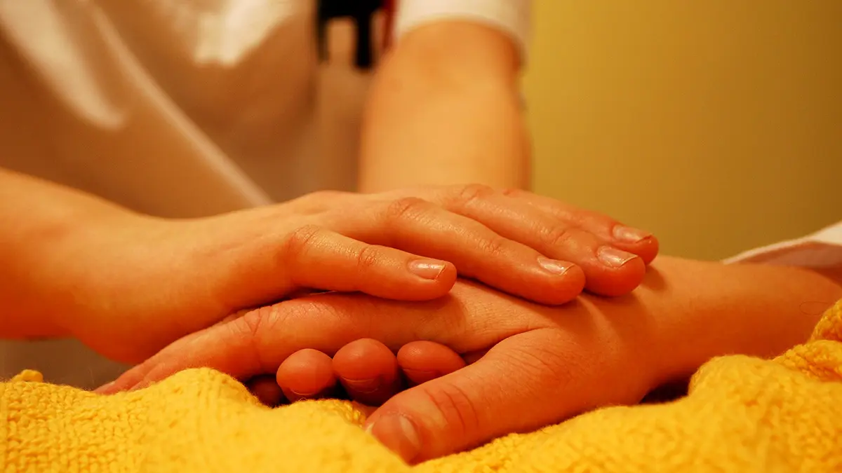 Hände einer pflegenden Person halten die Hand einer im Bett liegenden Person