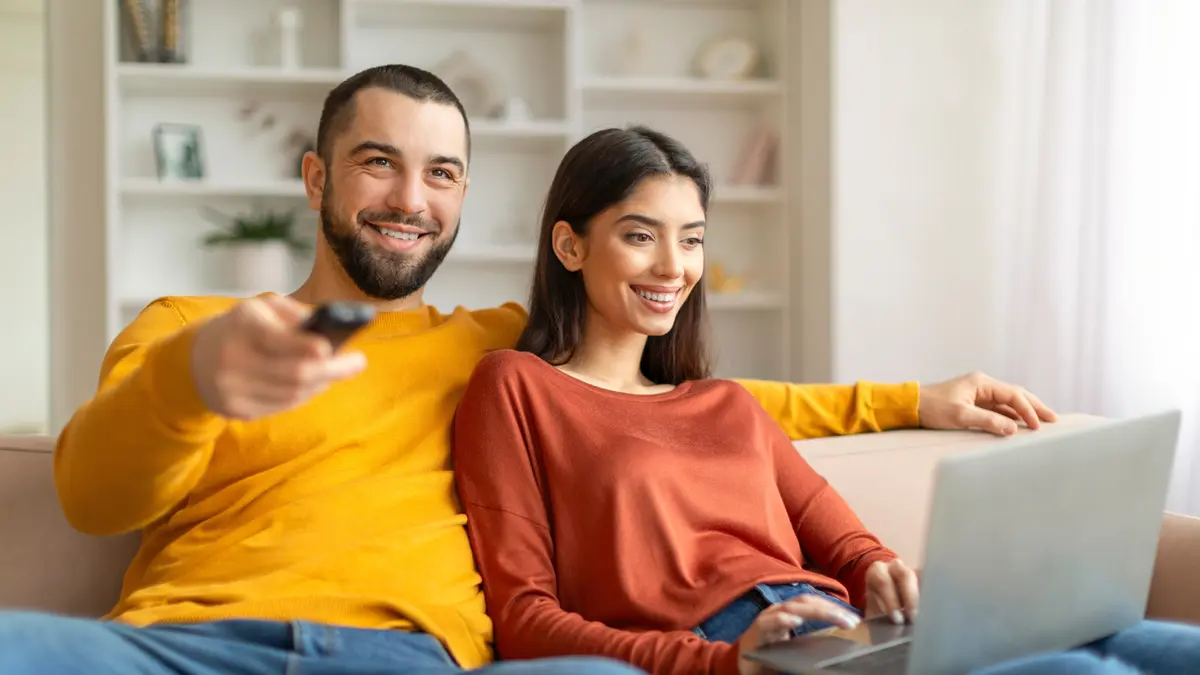 Mann und Frau sitzen auf dem Sofa, Frau hat Laptop auf dem Schoß, Mann hält Fernbedienung in der Hand und schaut TV. Beide lachen.