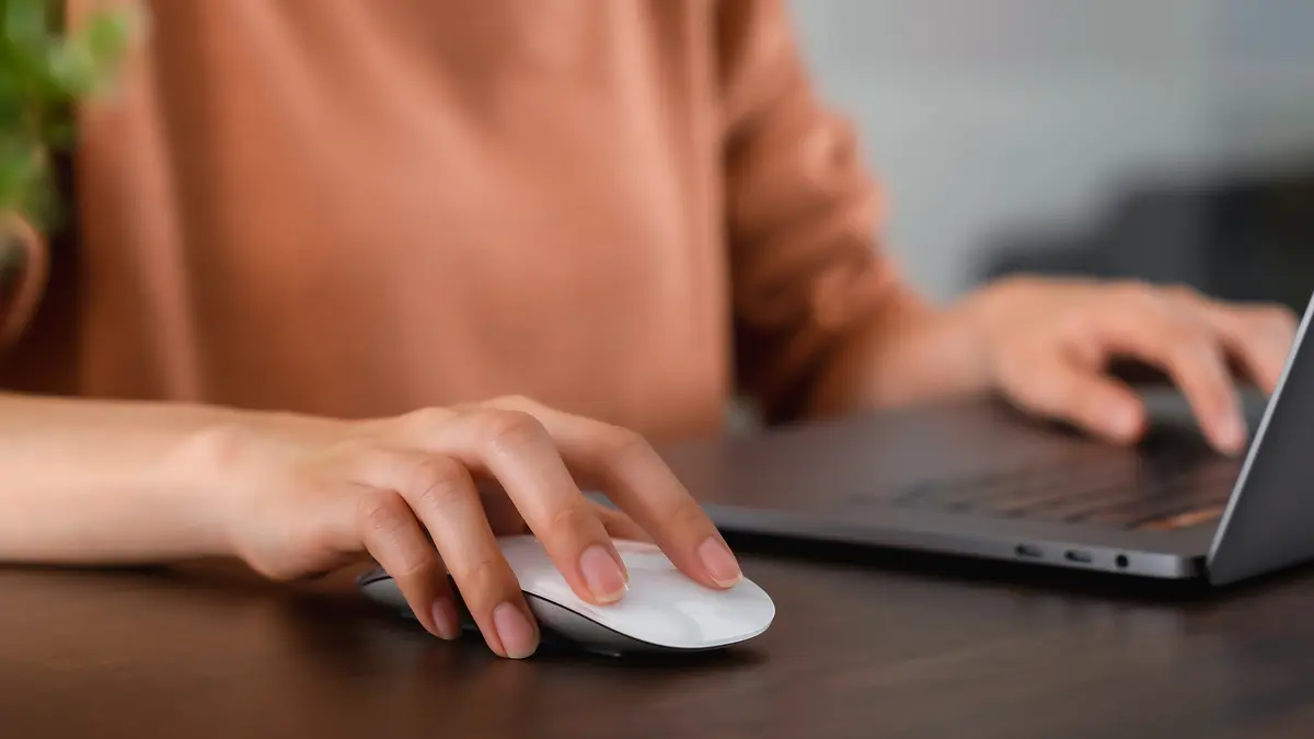 Eine Frau sitzt an ihrem Notebook und hält ihre Hand über eine Computermaus.