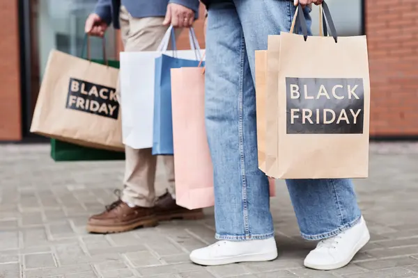 Menschen mit Papiertüten, auf denen die Worte Black Friday stehen, schlendern auf einer Straße. Es sind nur die Unterkörper zu sehen.