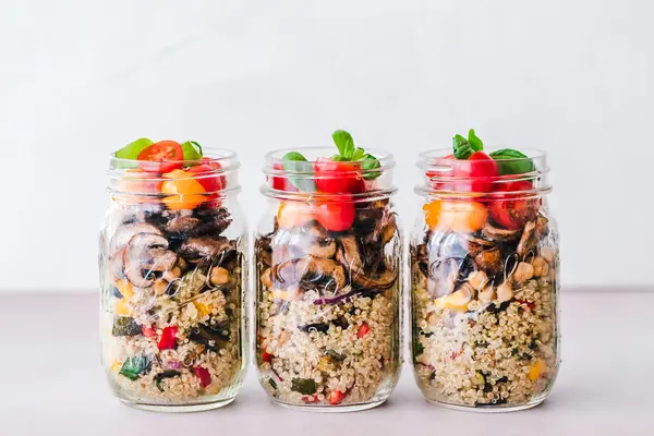 3 große Einmachgläser, welche jeweils mit Quinoa Salat (unterste Schicht), gebratenen Pilzen (mittlere Schicht) und halbierten Cherry-Tomaten und Basilikum (oberste Schicht) befüllt sind.