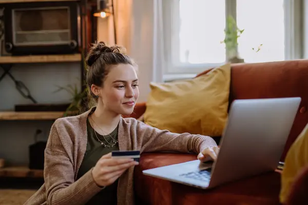 Eine Frau sitzt auf dem Boden vor ihrem Laptop, der auf einer ockerfarbenen Couch steht. Eine Hand greift an die Tastatur und mit der anderen Hand hält sie eine Kreditkarte. Sie lächelt.