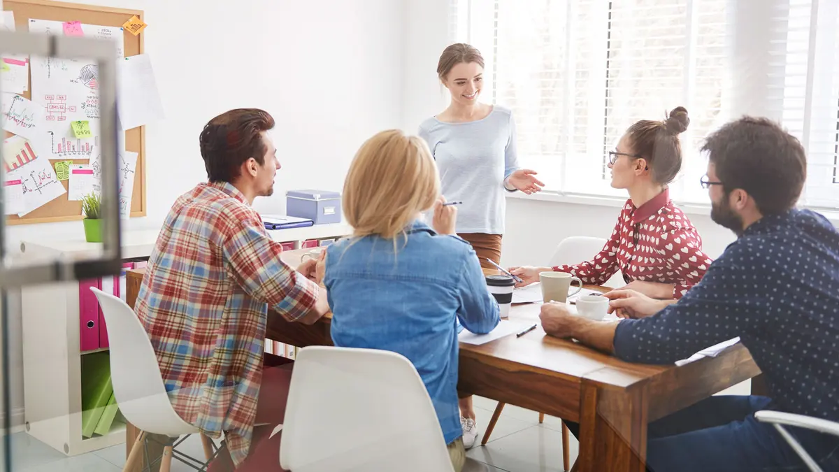 Ein Team sitzt um einen Besprechungstisch, eine Person steht und trägt etwas vor.