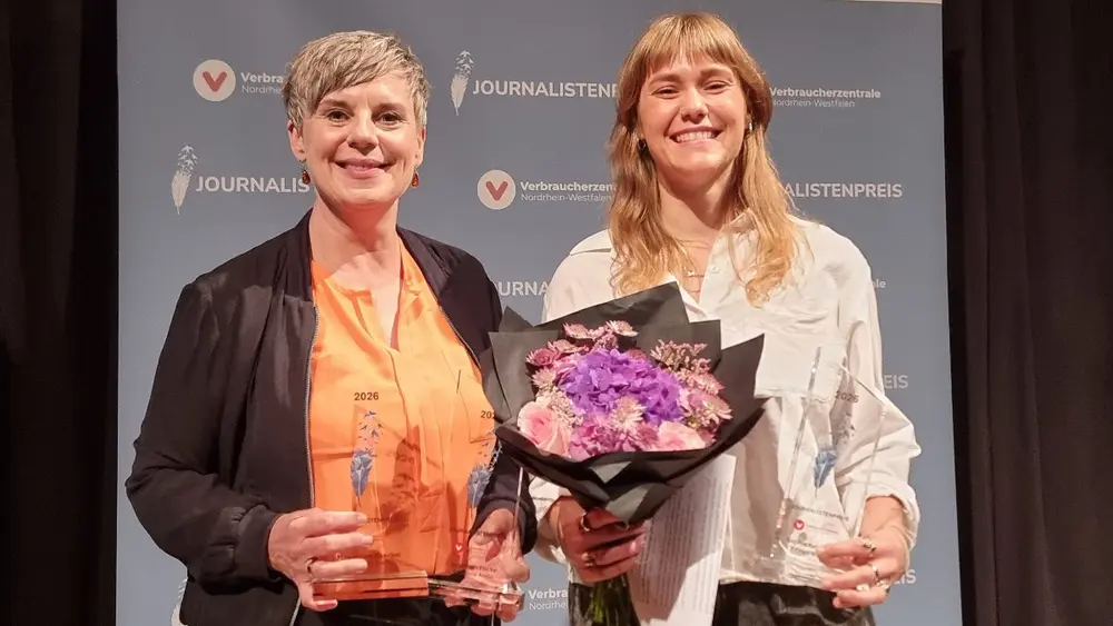 Zwei Frauen stehen nebeneinander und lächeln in die Kamera. Eine von ihnen hält einen Blumenstrauß und eine Trophäe in der Hand.