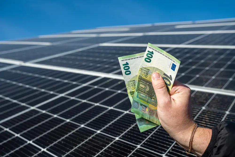 Eine männliche Hand hält zwei Hundert-Euro-Scheine in der Hand vor einer Photovoltaikanlage