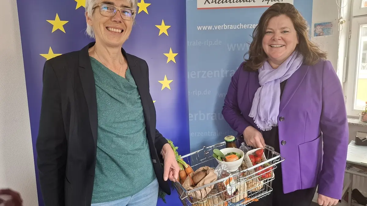 Zwei Frauen vor einem Roll-up mit der Europafahne. Sie halten einen Einkaufskorb zwischen sich.
