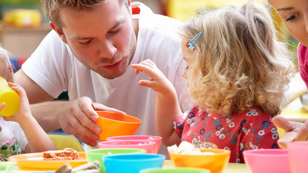 Ein Mann sitzt mit Kindern an einem Kindertisch mit bunten Schüsseln beim Essen