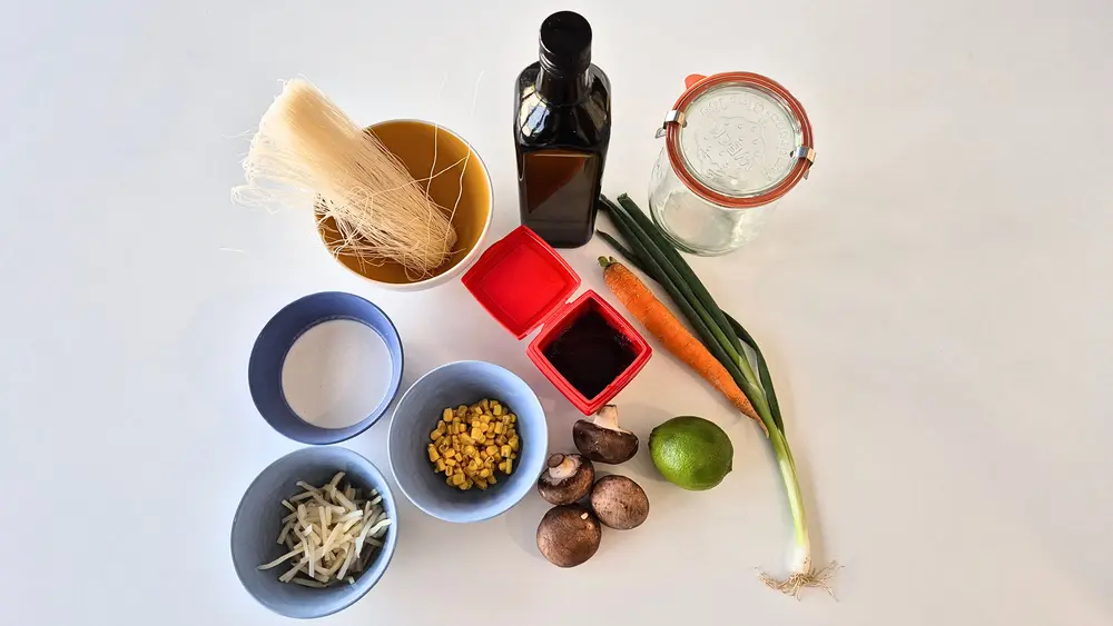 Zutaten auf einem weißen Hintergrund arrangiert: eine Schüssel mit Reisnudeln, eine Schüssel mit geraspelter Möhre, eine Schüssel mit Maiskörnern, eine Schüssel mit geschnittenen Frühlingszwiebeln, eine Flasche Olivenöl, ein Glas Kokosmilch, zwei kleine Behälter mit Miso-Paste und Gochujang, eine Limette, Pilze, eine Frühlingszwiebel mit Wurzel und ein Einmachglas zur Aufbewahrung.