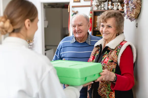 Eine junge Frau eines Lieferdienstes bringt zwei Senioren Essen in einer grünen Thermobox.