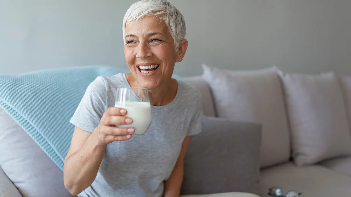 Lachende Frau mit grauen Haaren sitzt auf der Couch mit einem Glas Milch in der Hand