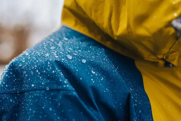 Jemand trägt eine blau-gelbe wasserabweisende Regenjacke mit abperlenden Regentropfen auf der Schulter