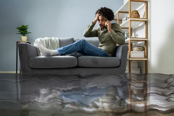 Ein Mann sitzt auf seiner Couch und hat ein Handy am Ohr. Seine Wohnung steht voller Wasser.