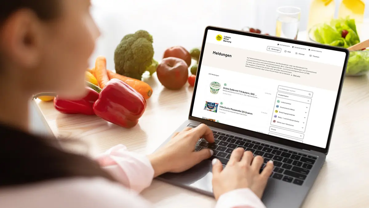Eine Frau sitzt mit Laptop am Küchentisch und schaut auf eine Internetseite auf dem Bildschirm.