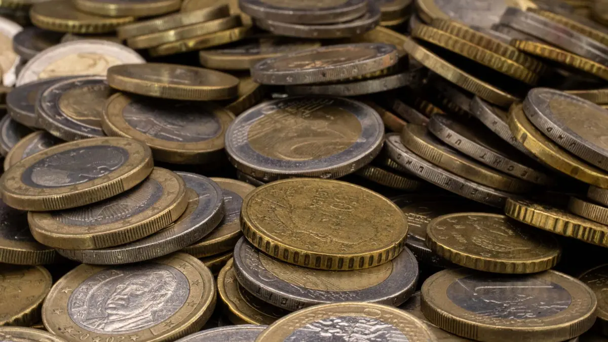 Eine Nahaufnahme zeigt einen großen, ungeordneten Haufen verschiedener Euro-Münzen, die übereinandergestapelt sind. Im Vordergrund sind deutlich 1-Euro- und 2-Euro-Münzen sowie goldfarbene Cent-Stücke (wahrscheinlich 50 Cent) zu erkennen. Die Münzen weisen Gebrauchsspuren und eine matte Textur auf, wobei das Licht die geprägten Ränder und Oberflächendetails hervorhebt.