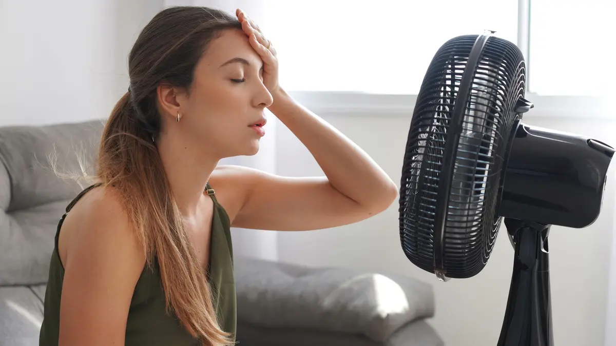 Eine junge Frau vor einem laufenden Ventilator, die zu Hause unter der Sommerhitze leidet.
