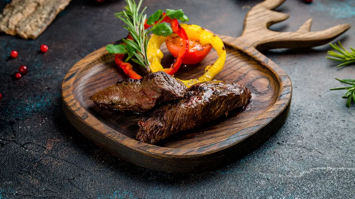 Ein Holzteller in Form eines Geweihs mit einem Stück gebratenem Wildfleisch drauf.