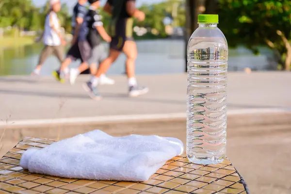 Im Vordergrund steht eine Wasserfasche aus PET, daneben liegt ein weißes Handtuch. Im Hintergrund joggt eine kleine Gruppe Läufer.