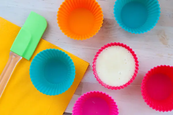 Bunte Silikonbackformen stehen auf einem Tisch, daneben liegen eine Silikonbackmatte und ein Silikonteigschaber. 