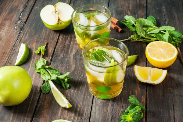 Zwei Gläser mit Alkoholfreien Getränken, daneben aufgeschnittene Äpfel, Zitronen, Kräuter und Zimtstangen