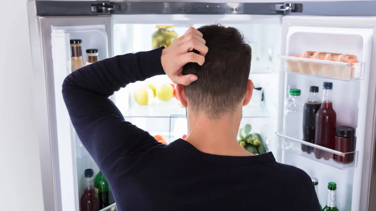 Ein Mann guckt in einen geöffneten Kühlschrank und kratzt sich dabei am Kopf.