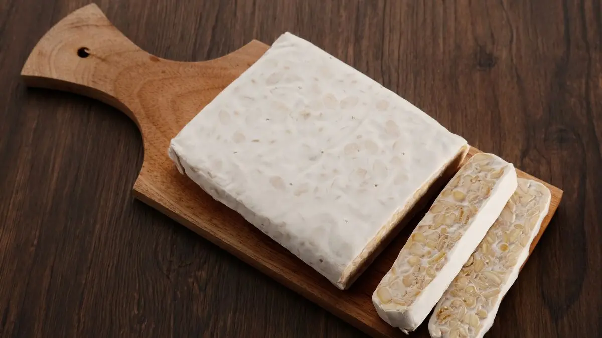 Eine Stück Tempeh liegt auch einem Holzbrett, zwei dünne Scheiben sind abgeschnitten.