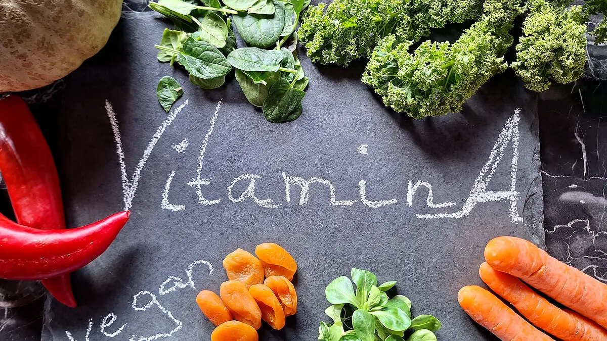 Verschiedene Vitamin A-haltige Gemüse wie Spinat und Möhren liegen auf einer schwarzen Schiefertafel auf der "Vitamin A" und "vegan" steht.