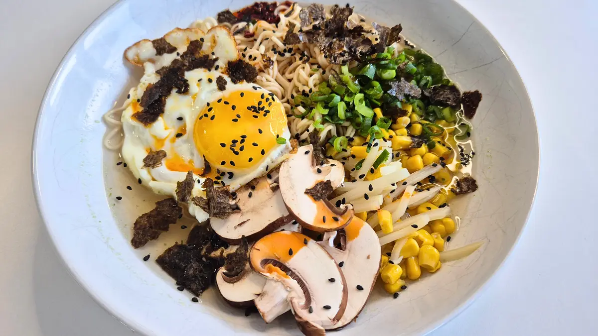 Ein weißer Teller mit upgegradeten Instant-Nudeln: cremige Nudeln mit Spiegelei (dottergelb, mit Sesam bestreut), gebratenen Pilzscheiben, frischen Frühlingszwiebelringen, Maiskörnern und Sojasprossen. Alles garniert mit knusprigen Algenblättern (Nori) und Sesamsamen.