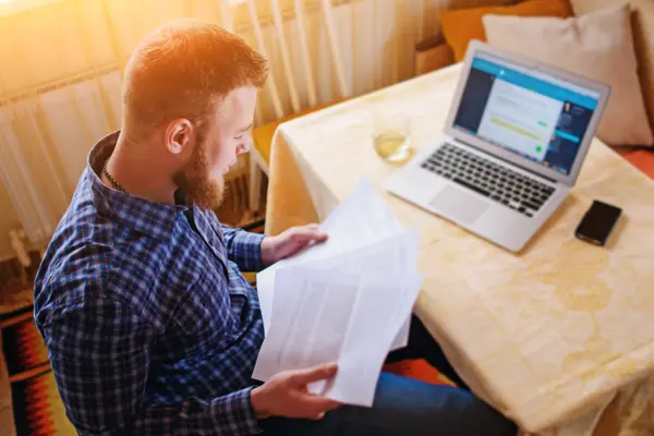 Ein Mann sitzt in seiner Wohnung am Laptop und studiert ausgedruckte Unterlagen