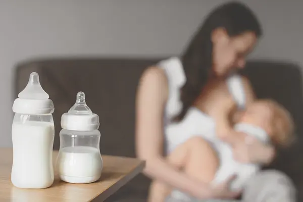 Zwei Babyflaschen auf einem Tisch, Mutter stillt Baby im Hintergrund