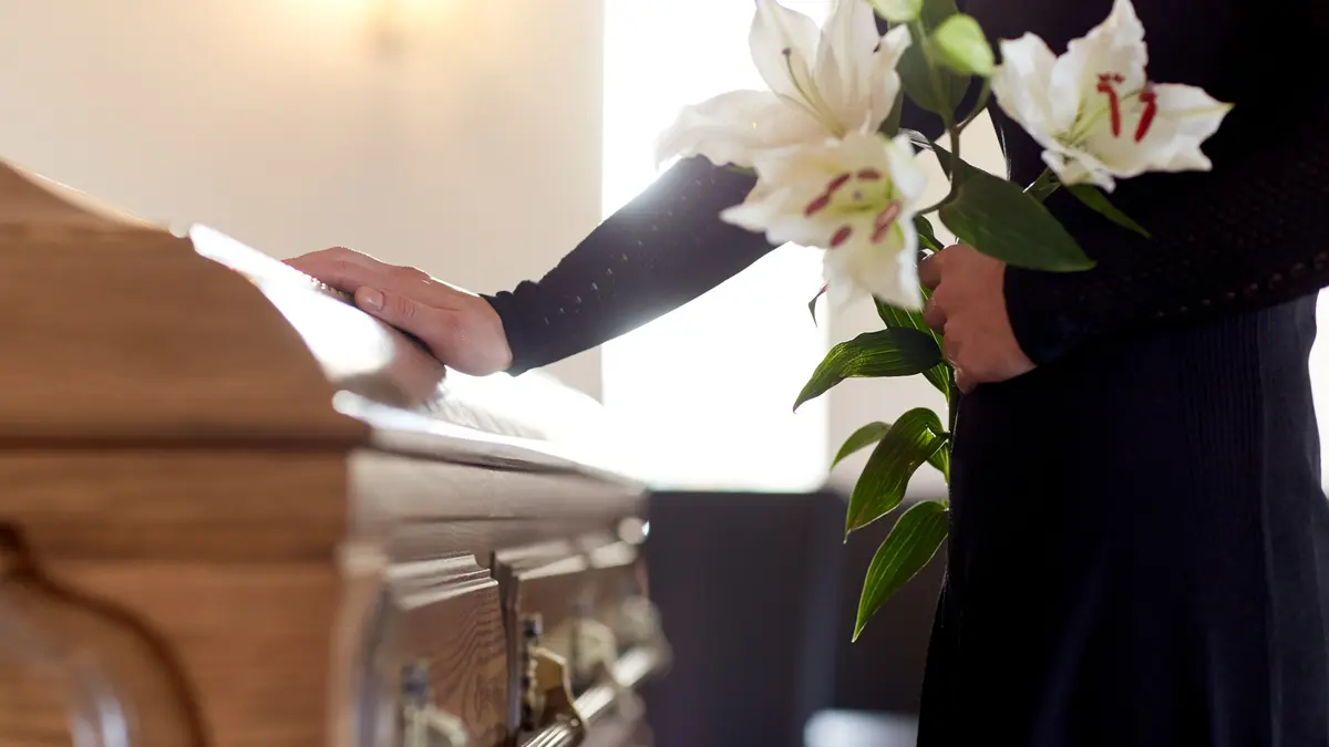 Eine Frau steht mit Blumen in der Hand an einem Sarg.