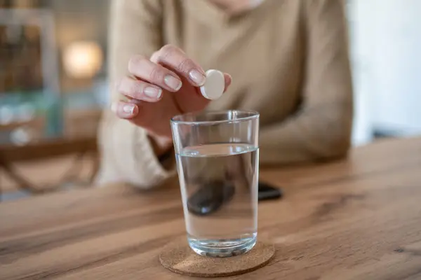 Eine Frauenhand hält eine weiße Tablette über ein Wasserglas