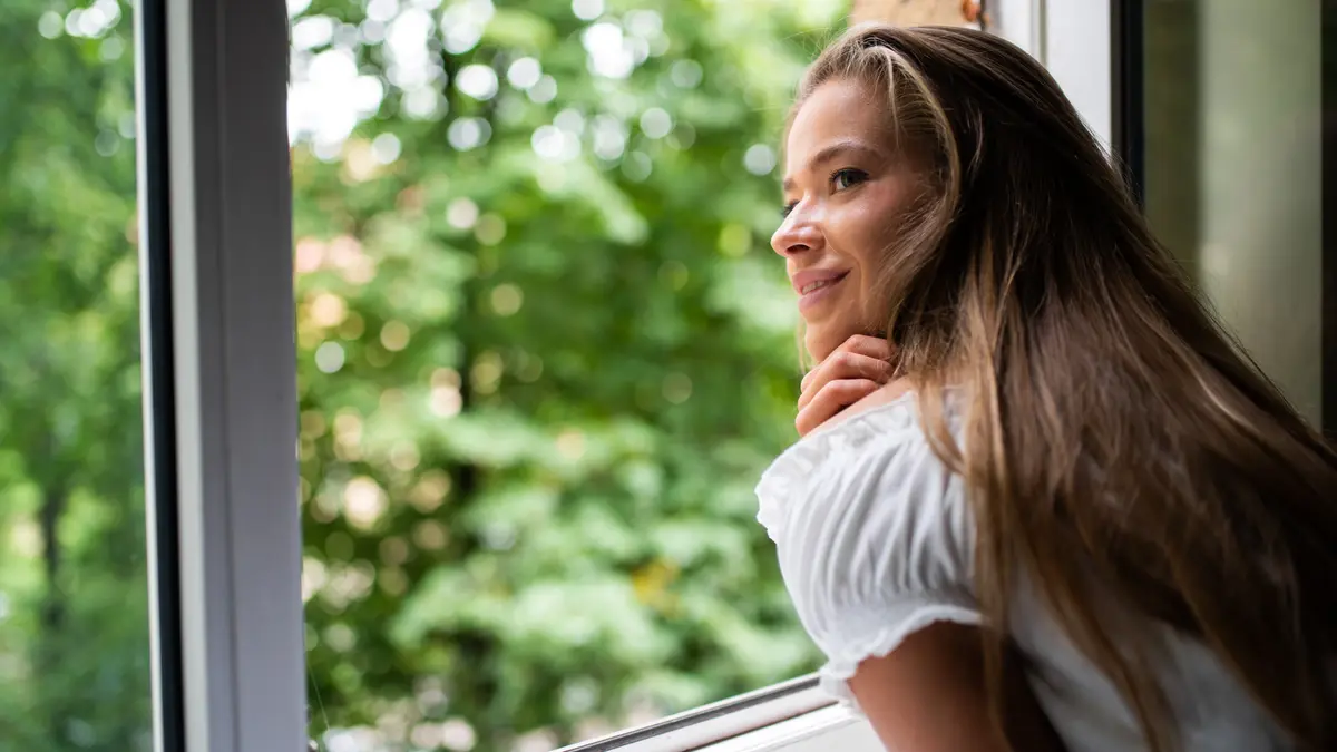 Eine junge Frau lehnt sich auf die Fensterbank eines offenen Fensters und blickt nach draußen ins Grüne.