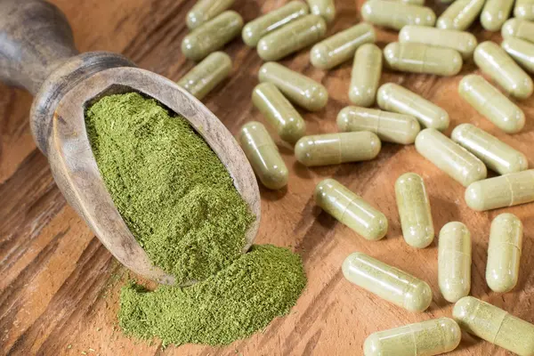 grünes Moringa-Pulver in einer Holzschaufel, dazu verstreut liegende grüne Kapseln