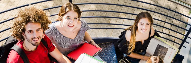 Vier junge Erwachsene sitzen mit Büchern auf einer Wendeltreppe und schauen nach oben in die Kamera 