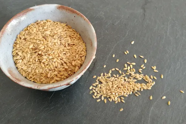Eine helle Schüssel mit Leinsamen steht auf einem Tisch, neben der Schüssel liegen ebenfalls einige Leinsamenkörner
