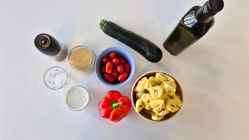 Von oben fotografierte Zutaten für ein schnelles Tortellini-Rezept: Tortellini, Paprika, Zucchini, Kirschtomaten, Sahne, Gewürze, Parmesan und Olivenöl auf einem hellen Tisch arrangiert.