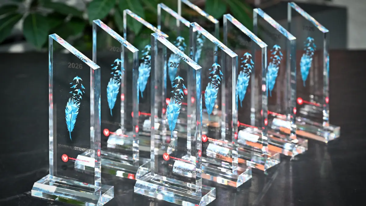 Transparente Trophäen aus Plexiglas stehen aufgereiht auf einem Tisch, jede trägt das Bild einer blauen Feder, aus der am oberen Bereich Vögel aufsteigen.
