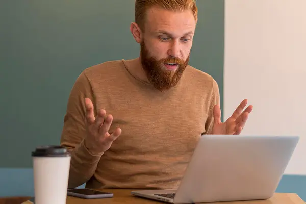 Ein Mann sitzt vor einem Laptop und hebt verärgert die Hände