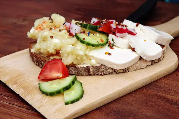 ein belegtes Brot mit Käse, Gurke und Tomate auf einem Brettchen