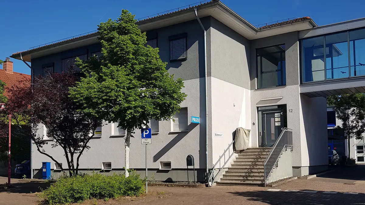 Ansicht eines grau gestrichenen Hauses mit zwei Bäumen und einen Parkplatz für beeinträchtigte Personen davor. Treppenaufgang zur Eingangstür mit elektrisch fahrbarer Bühne. Vlauer Himmel im Hintergrund.
