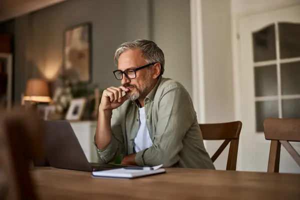 Ein Mann mittleren Alters mit Brille sitzt am Esstisch vorm Laptop. Vor ihm liegt ein Notizblock mit Stift. Er hat einen Arm auf den Tisch gestützt und hält die Hand nachdenklich an sein Kinn. Er lächelt vage. 